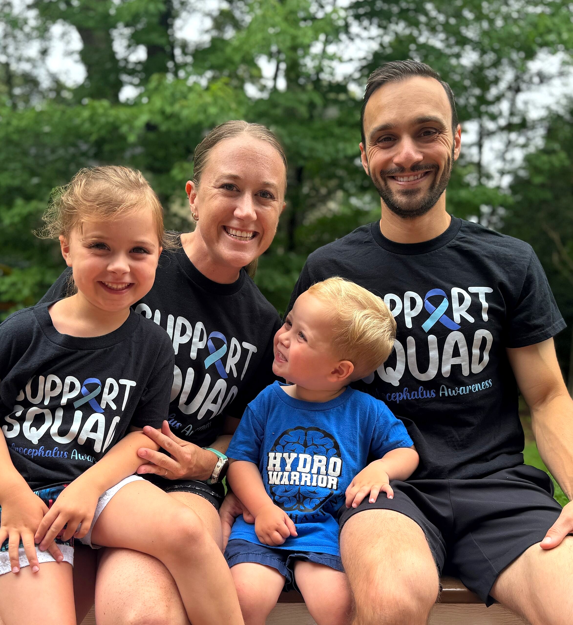 A mother, father, daughter and toddler son in matching Support Squad hydrocephalus awareness shirts, the boy looking up at his dad