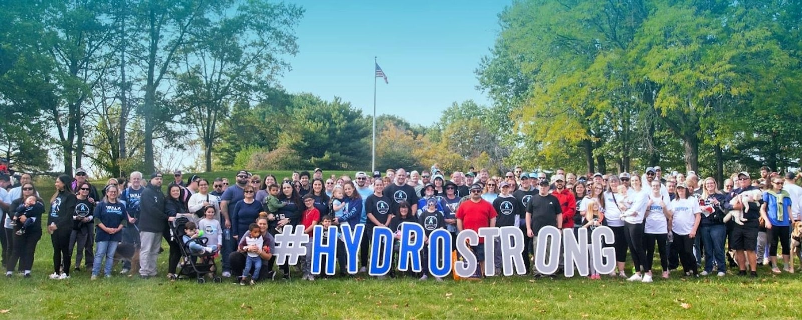 Hundreds of WALK participants in blue shirts behind giant #HYDROSTRONG letters on a sunny lawn